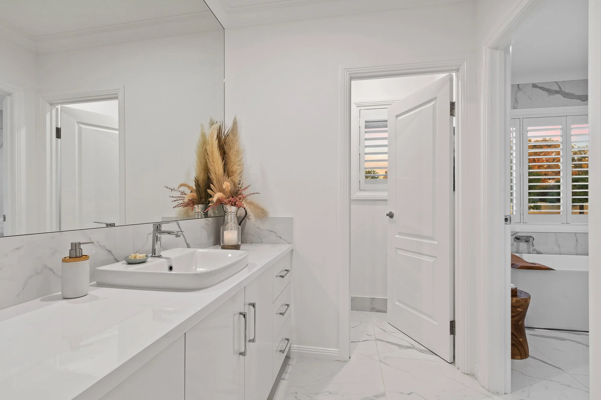 Modern bathroom with white marble countertop, vessel sink, and decorative vase with dried pampas grass and flowers, large mirror, and white painted walls.