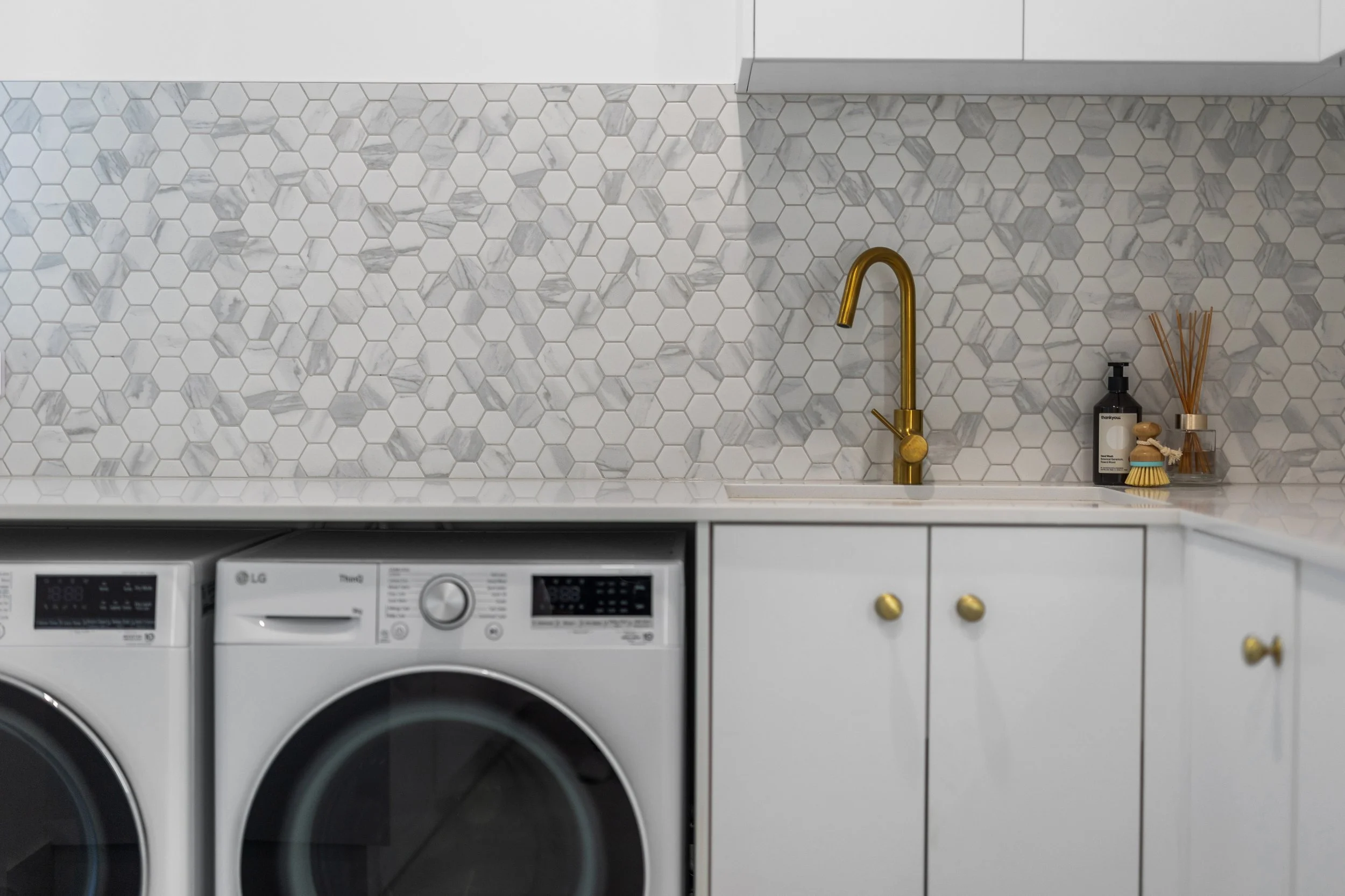 Laundry room with white hexagonal-tile backsplash, gold faucet, white cabinetry with gold knobs, behind a washing machine, with soap dispenser, brush, and reed diffuser on counter.
