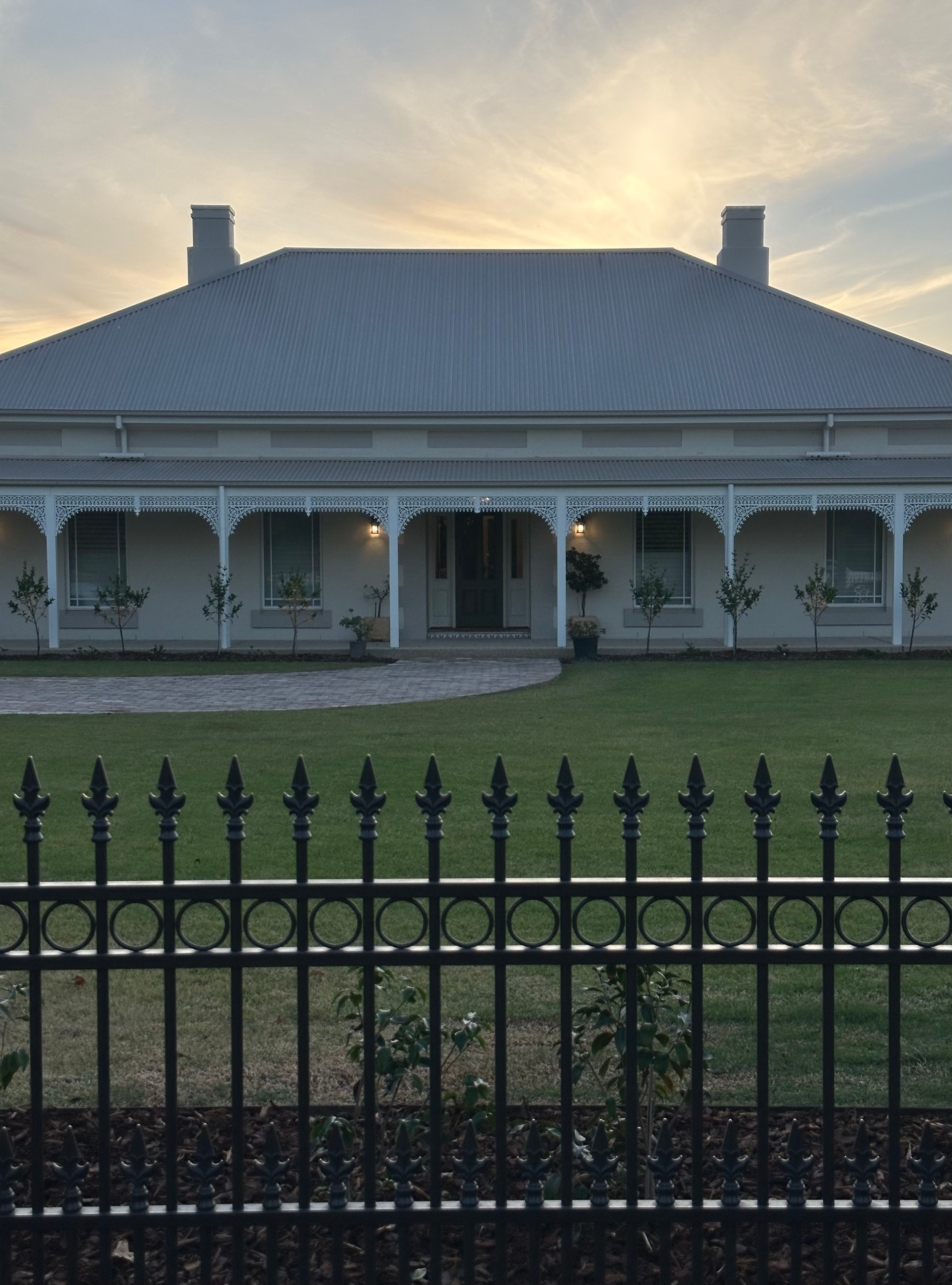 A large house with a metal roof, a front porch with decorative trim, and a fence in the foreground. The house is painted white with large windows and small trees along the front. The sky is partly cloudy with the sun setting or rising in the background.