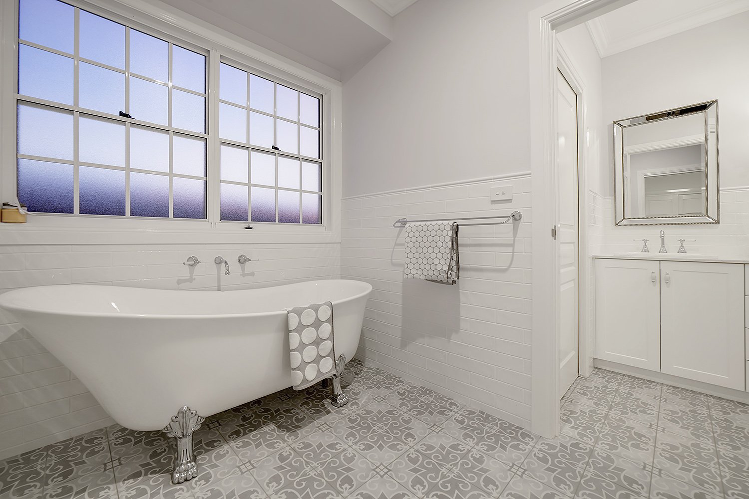 A bright bathroom with a white clawfoot bathtub, large frosted window, white paneled walls, and patterned floor tiles. There is a towel hanging on a wall-mounted towel rack and a mirror above a white vanity cabinet.