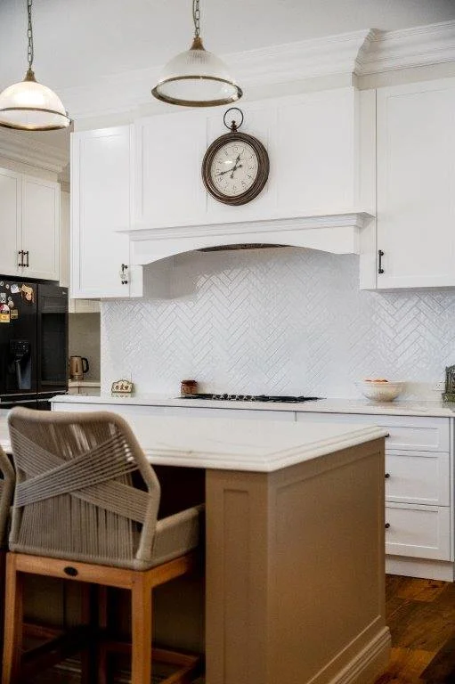 A cozy kitchen with white cabinets, a white countertop, and a herringbone tile backsplash. There is a wall clock above the range hood, pendant lighting, a dining chair at a breakfast bar, and a black refrigerator on the left side.