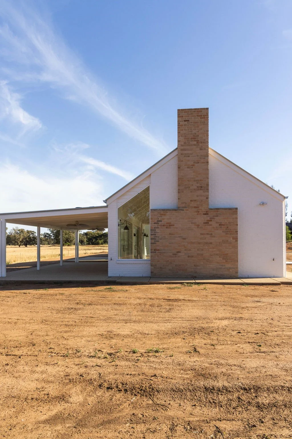 A modern house with a white exterior, large glass window, and prominent brick chimney against a blue sky with scattered clouds. The house is in a rural setting with dirt ground and open fields in the background.