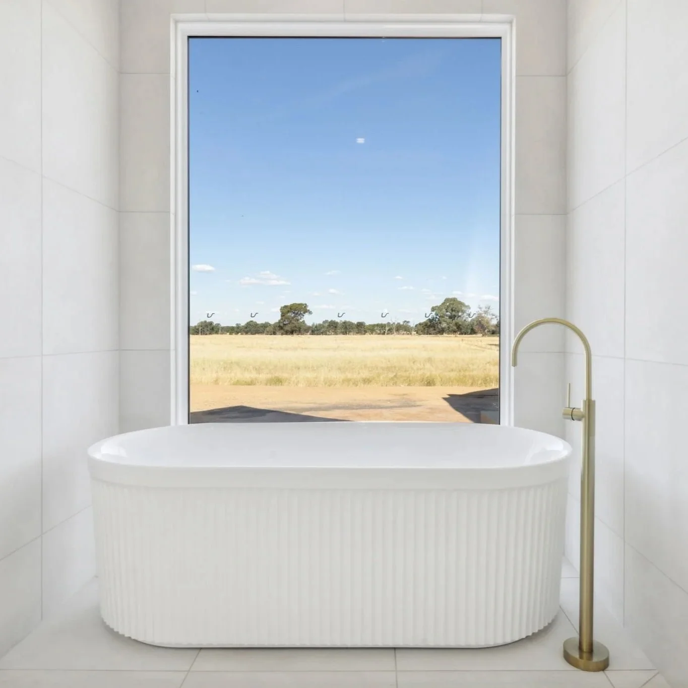 White bathtub in a bathroom with a large window overlooking a field and trees under a blue sky.
