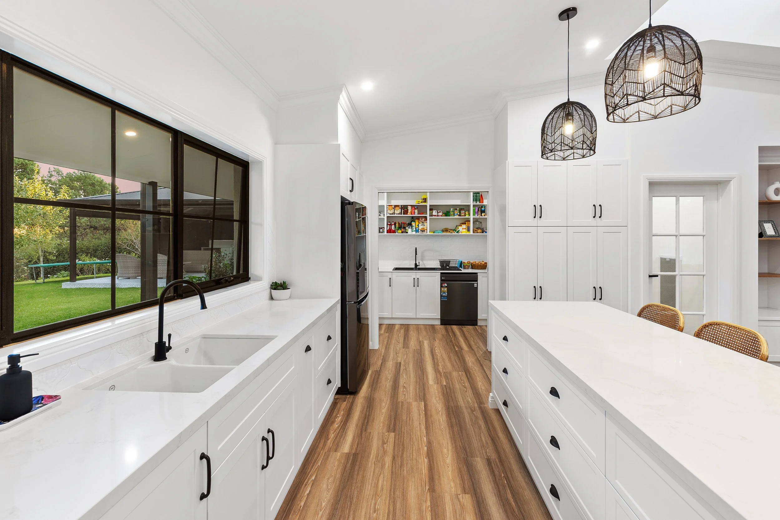 Modern kitchen with white cabinets, large island with drawers, black pendant lights, black faucet, stainless steel refrigerator, open shelves with food, and large window showing backyard