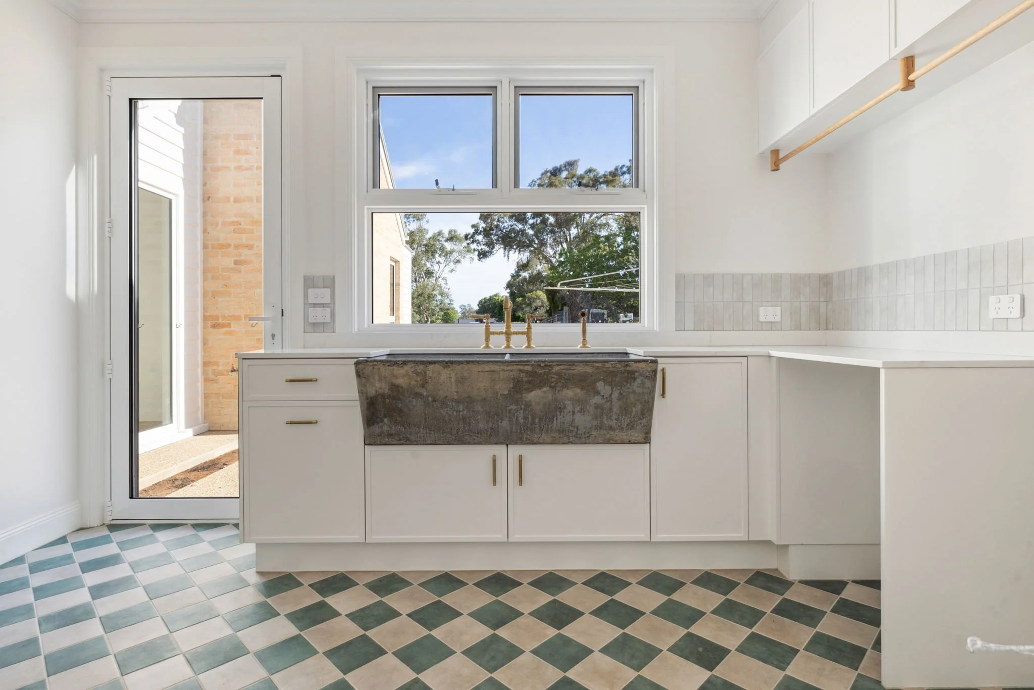 Kitchen with white cabinets, a large window, and a farmhouse sink with brass fixtures, with a door leading outside, checkered floor tiles, and a countertop on the right.