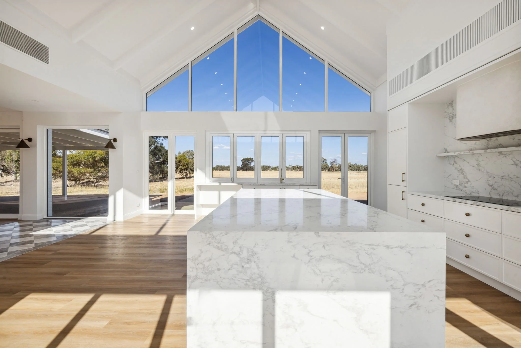 Modern kitchen with white marble island and cabinetry, large windows and glass doors leading to an outdoor view of fields and trees, high vaulted ceiling, hardwood floors, and natural sunlight.