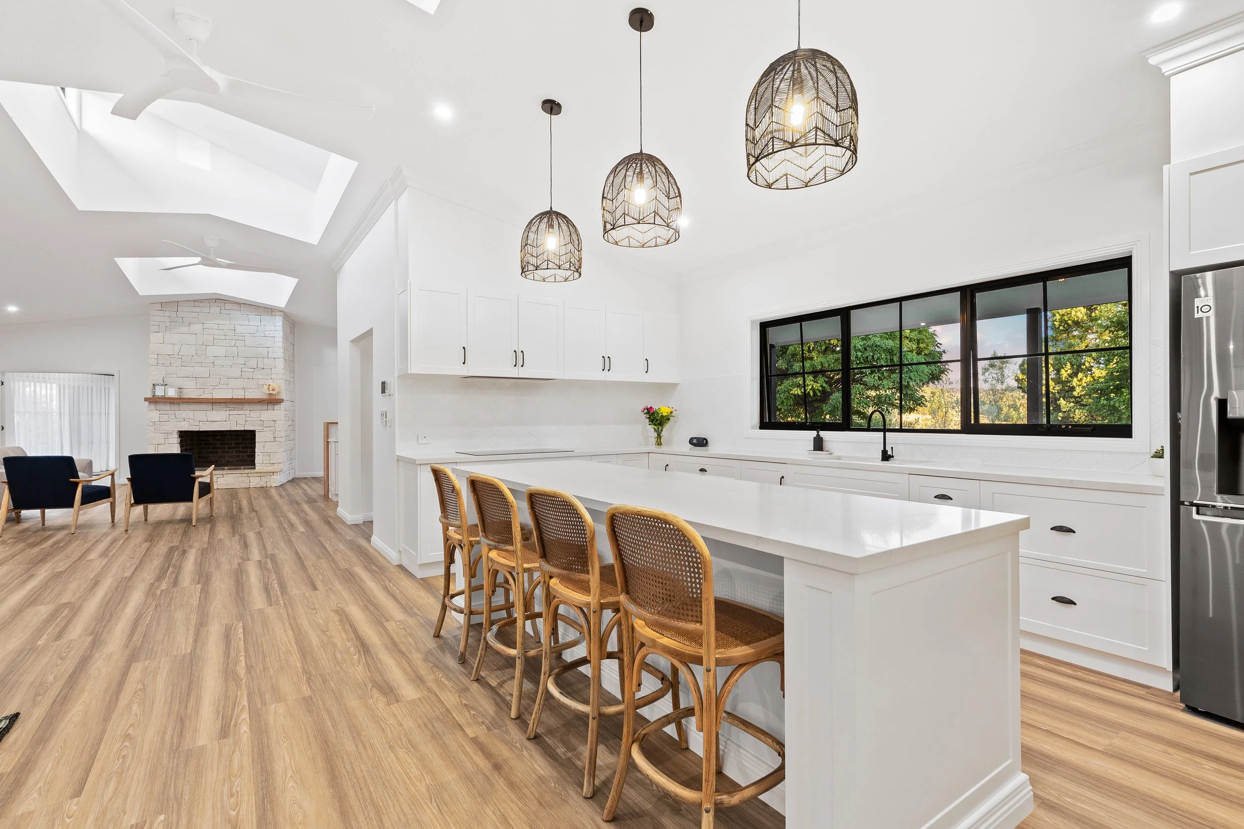 Modern kitchen with white cabinets, island counter, pendant lights, black-framed window, and wooden bar stools, with a view of a green outdoor area.