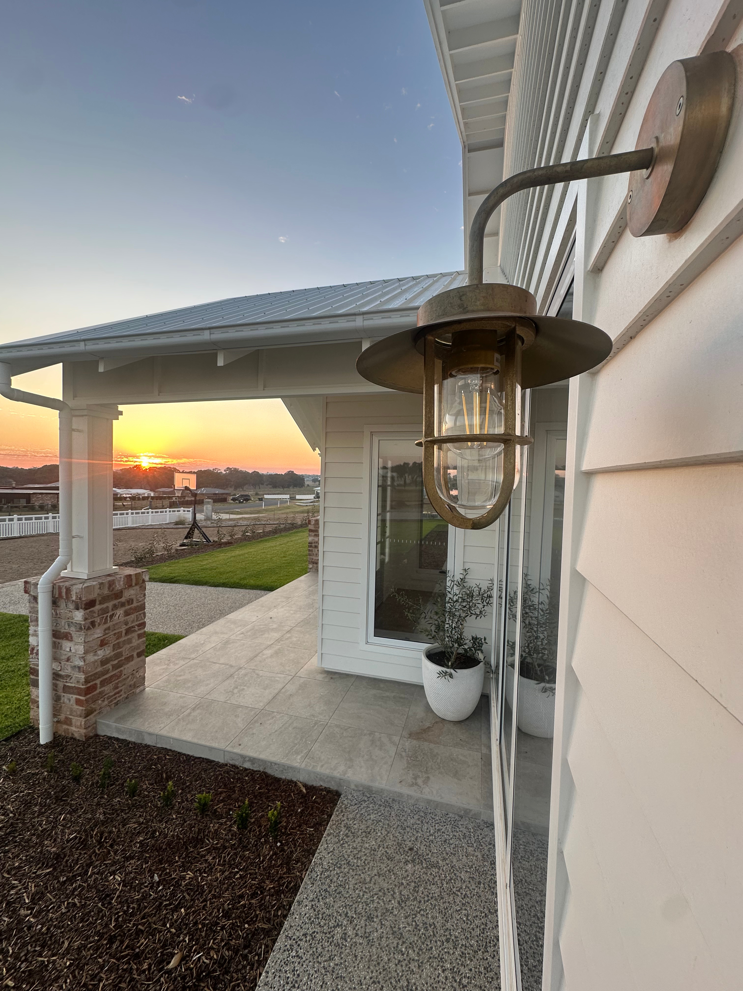 Exterior of a house with a porch, white siding, and a metal lantern-style light fixture near the door, with a sunset in the background.