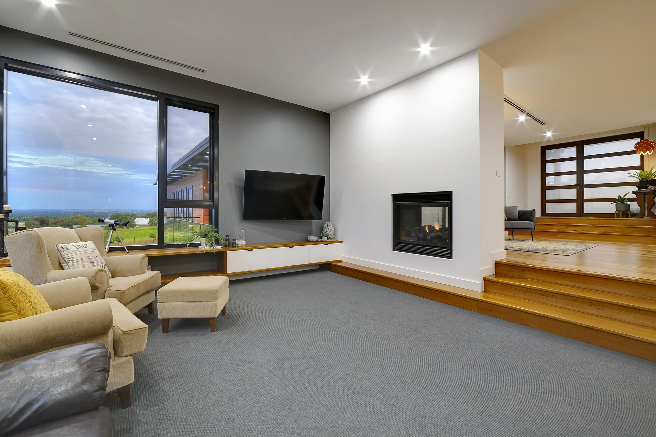 Living room with large window showing view of fields and sky, beige armchair with matching ottoman, dark gray accent wall with mounted TV, modern fireplace, wooden flooring with step leading to another sitting area with a gray chair and plants.