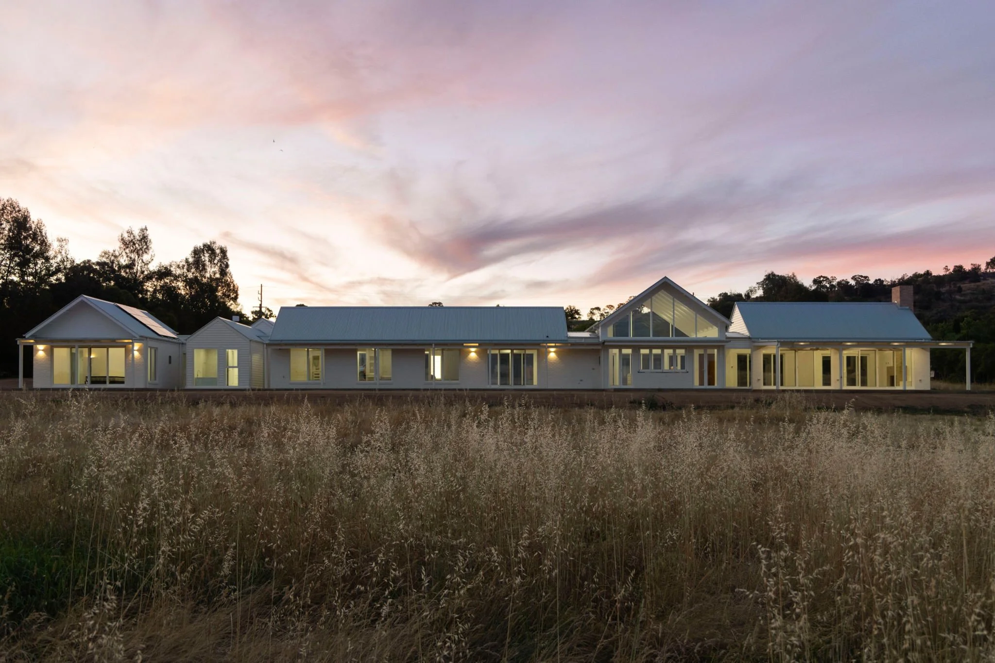 Modern single-story house with large windows and a metal roof, illuminated from inside, situated in a field during sunset with a pink and purple sky.