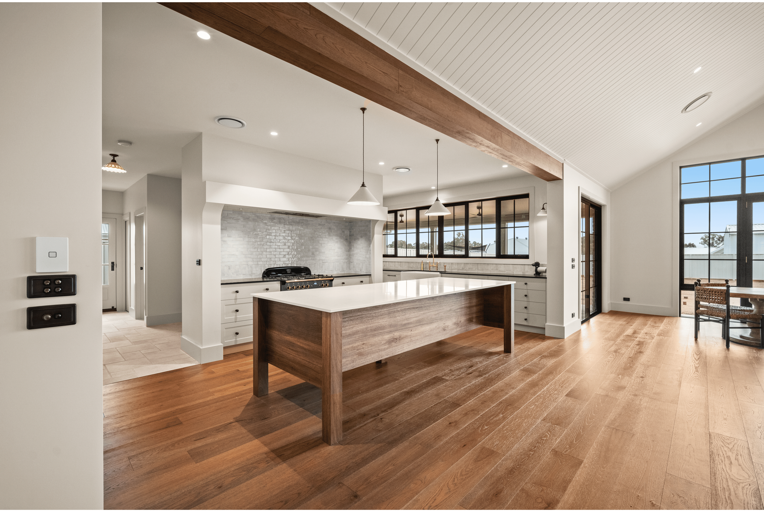 Modern kitchen with white marble island and cabinetry, large windows and glass doors leading to an outdoor view of fields and trees, high vaulted ceiling, hardwood floors, and natural sunlight.