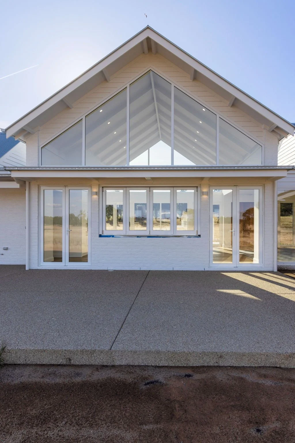 Modern house with large glass windows and doors, white exterior, and a sloped roof. The house has a spacious outdoor area with a concrete patio.