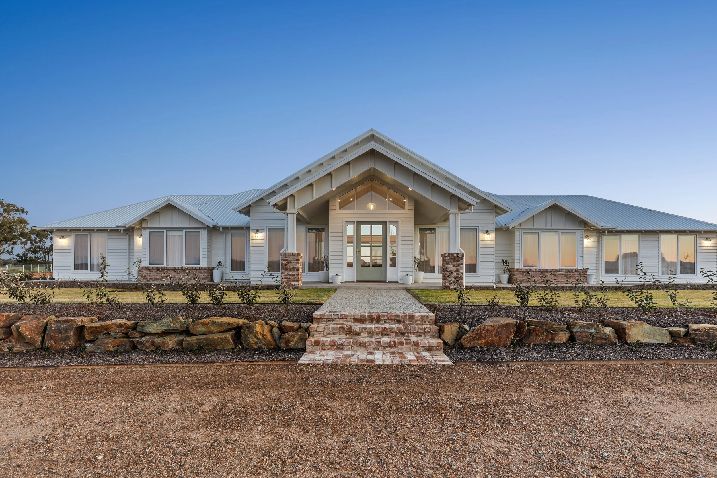 Front view of a modern, single-story house with white siding, large windows, and a covered porch, with a stone pathway and landscaped yard.