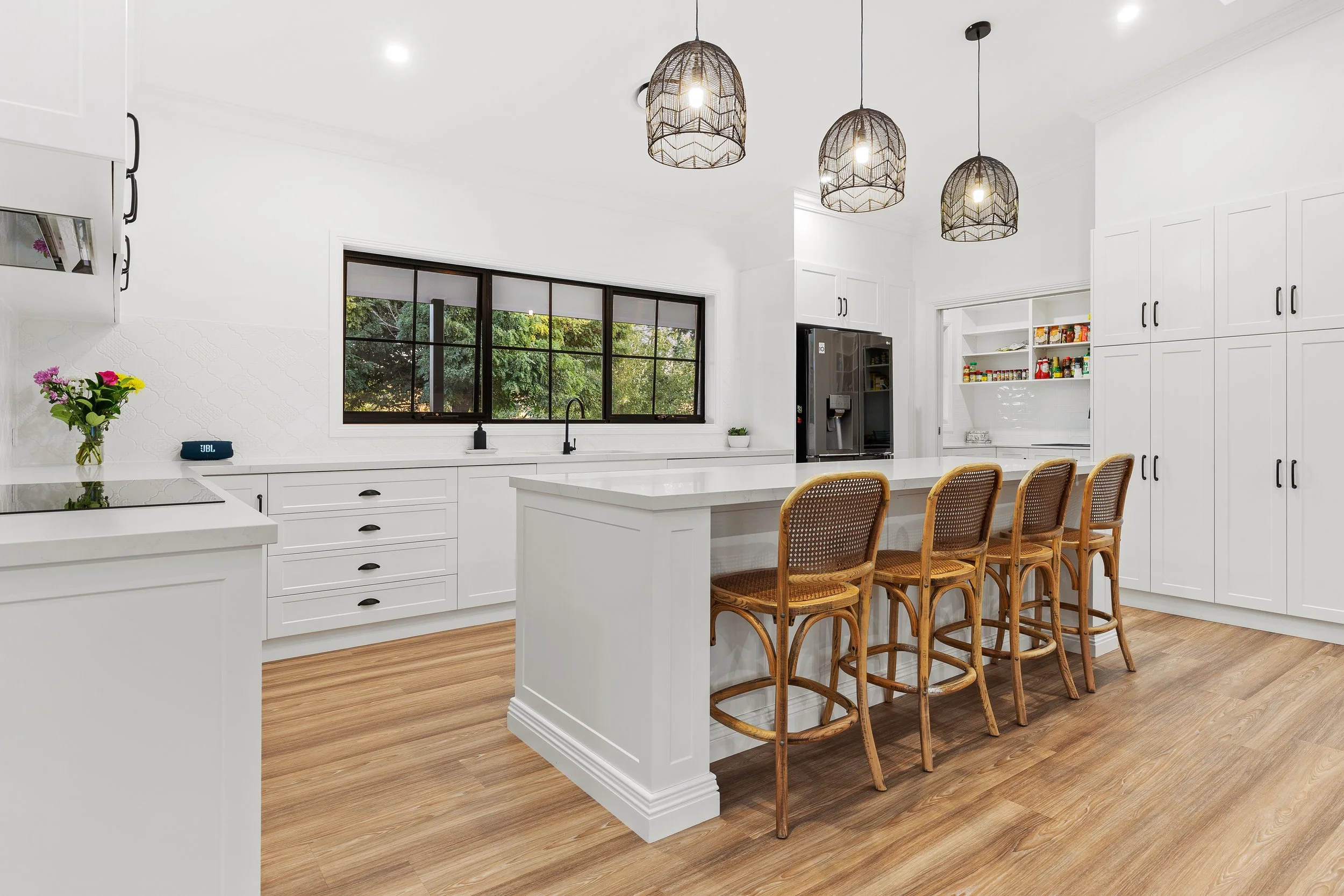 Modern white kitchen with island and four wooden barstools, black window, black appliances, hanging farmhouse light fixtures, light wood flooring, and open pantry with spices.