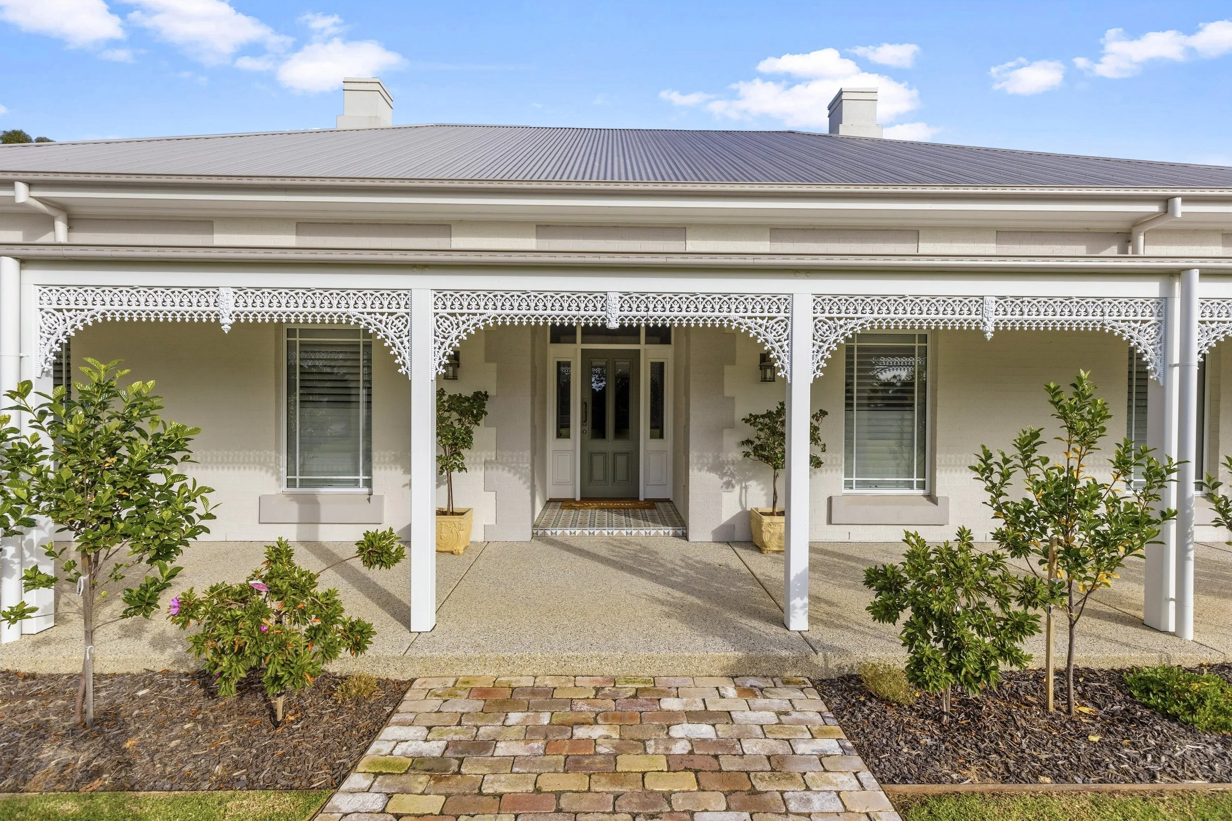 A large house with a metal roof, a front porch with decorative trim, and a fence in the foreground. The house is painted white with large windows and small trees along the front. The sky is partly cloudy with the sun setting or rising in the background.