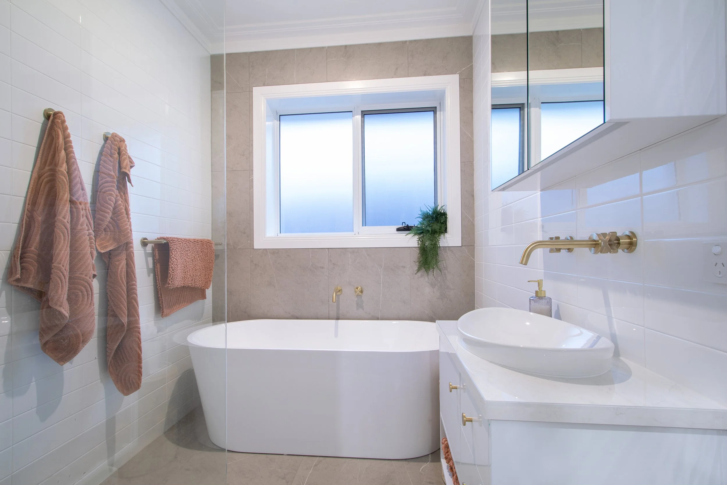 Modern bathroom with a freestanding bathtub beneath a frosted window, white tiled walls, a vanity with a white sink, gold fixtures, and pink towels hanging on hooks.