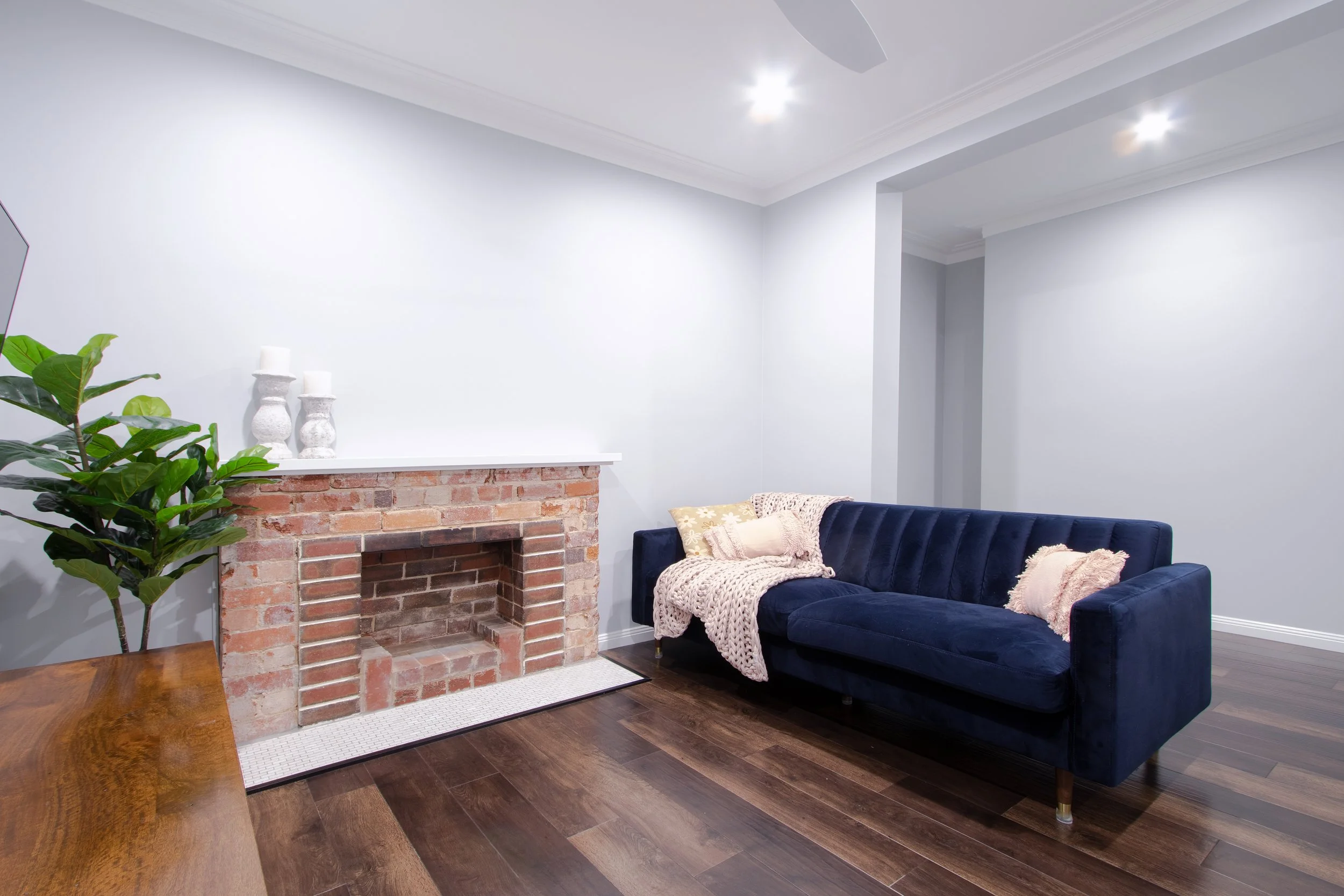 Living room with a blue velvet couch, a brick fireplace with a white mantel, a large green plant in the corner, and wooden flooring.