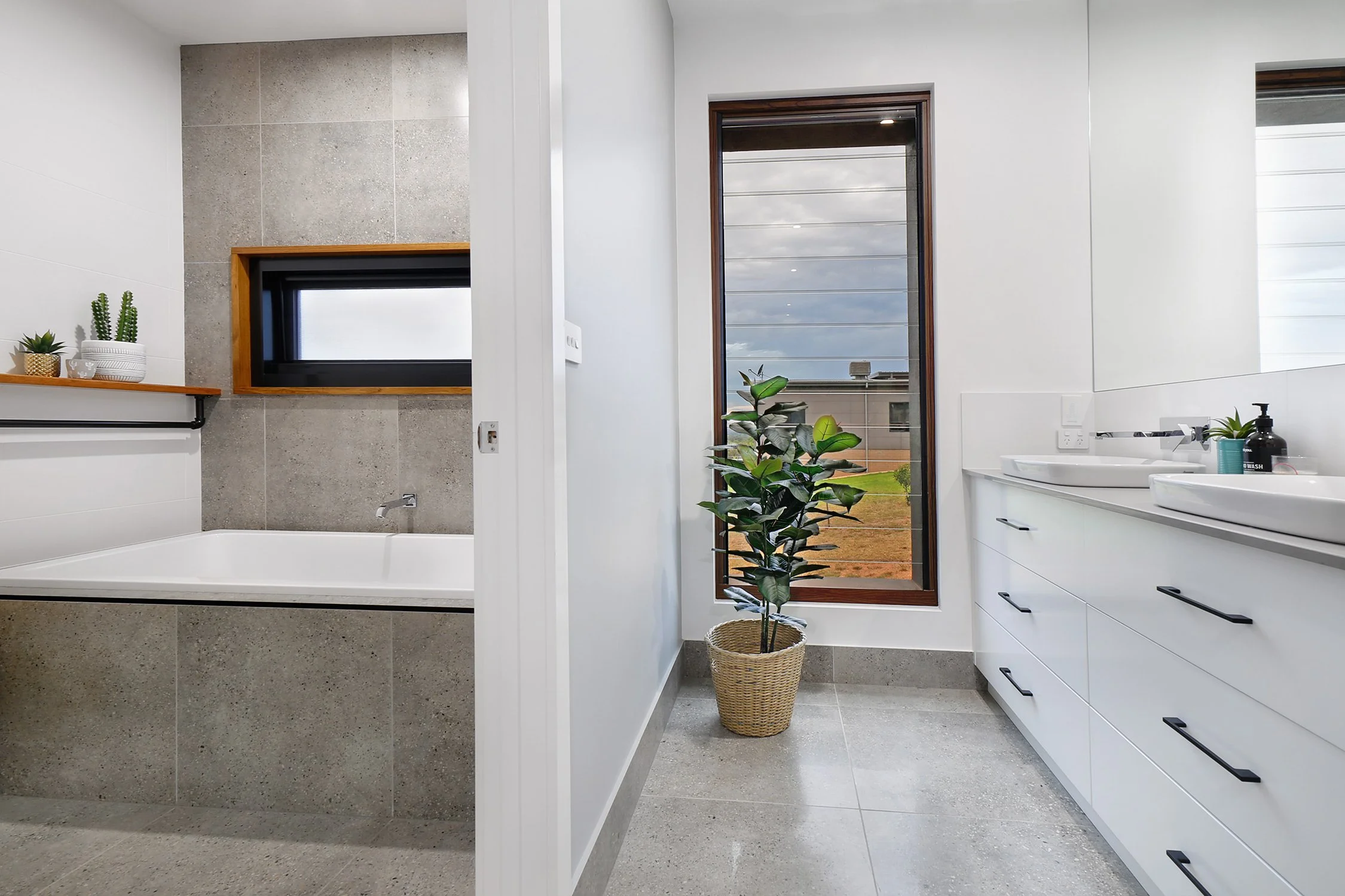 Modern bathroom with a bathtub, small window, tall mirror, two sinks, white cabinetry, a large window showing a backyard, and a potted plant.