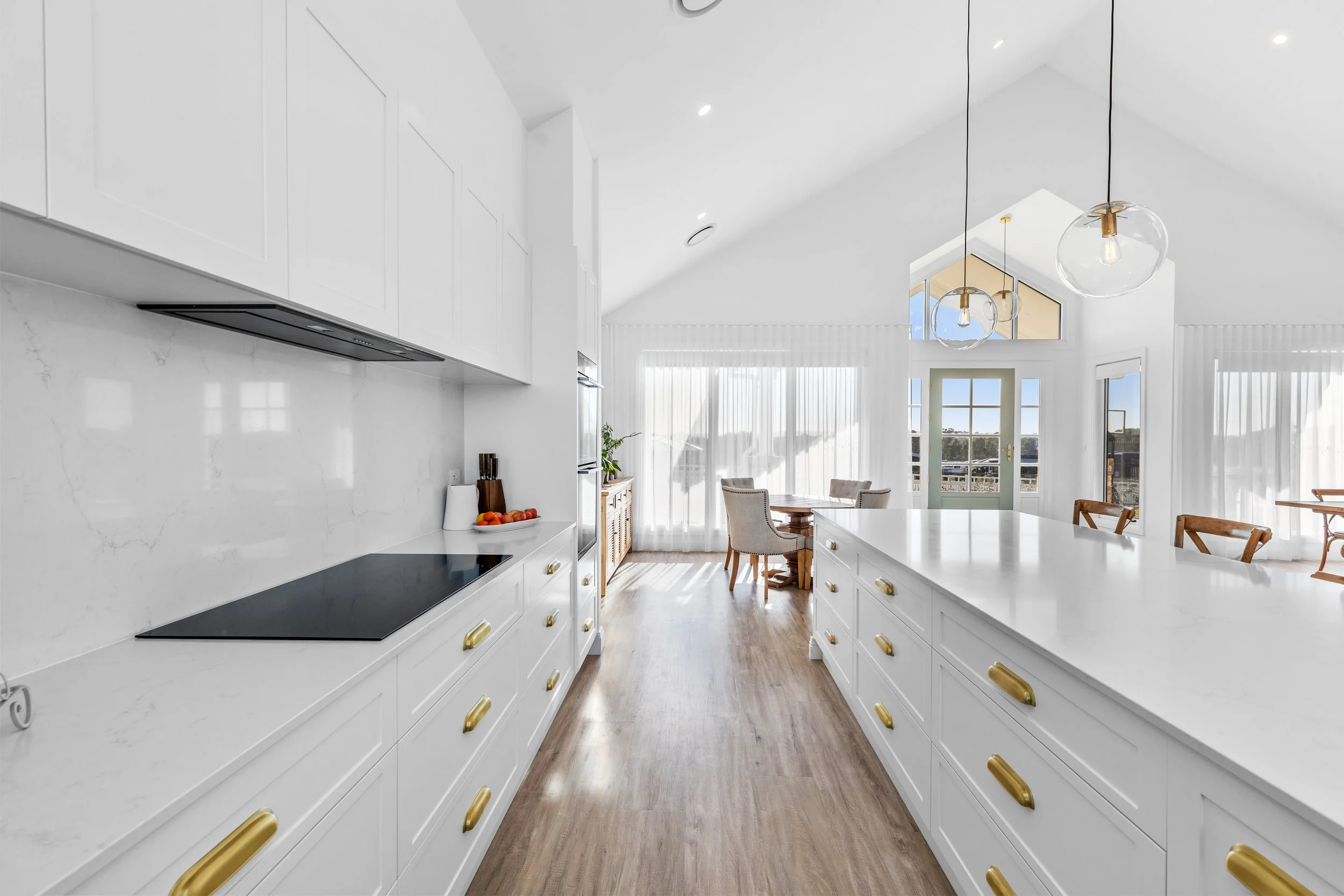 Bright, modern kitchen with white cabinetry, marble backsplash, and gold hardware, open to a dining area with windows and glass door.
