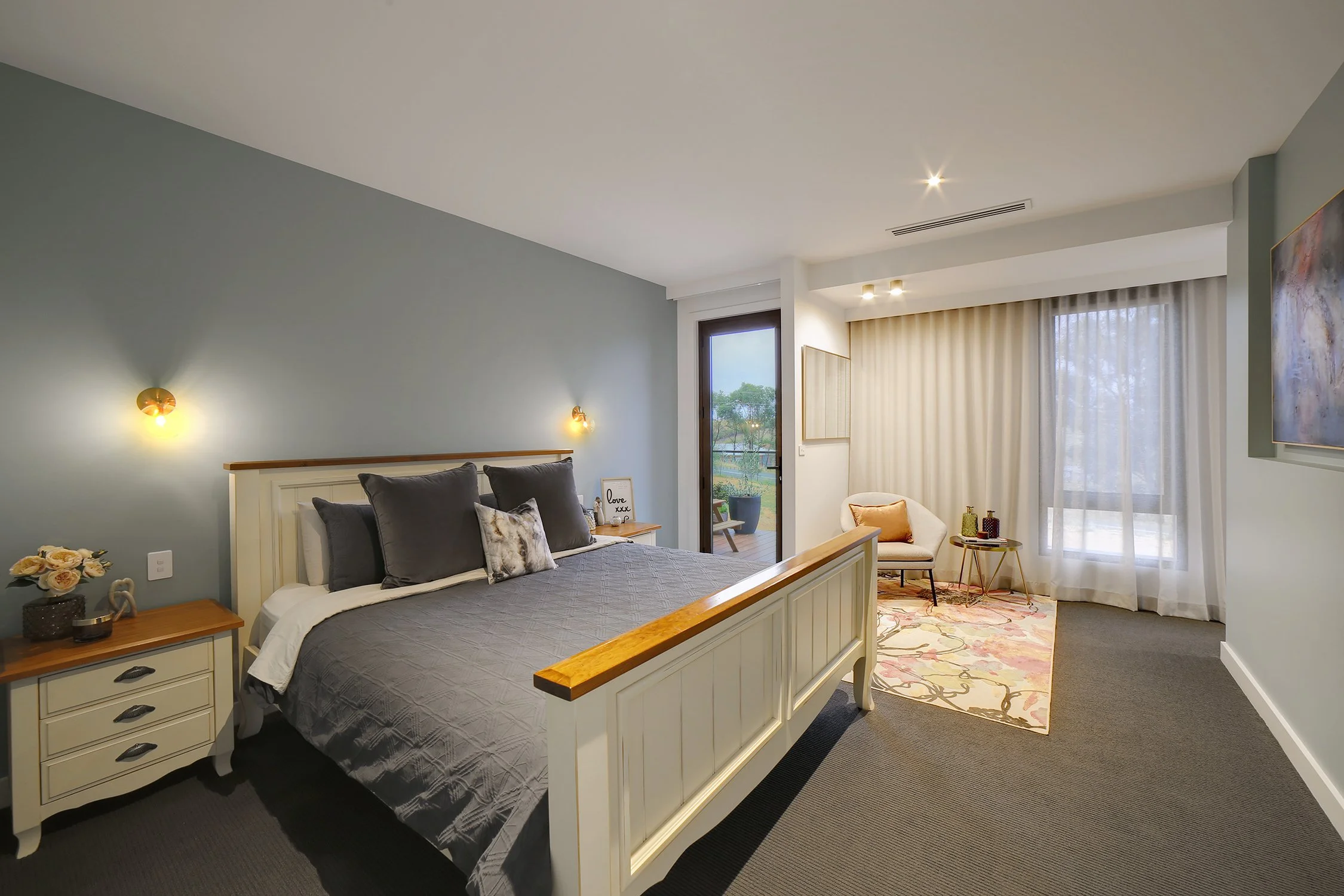 A modern bedroom with a large bed, gray bedding, and multiple pillows, adjacent to a balcony with outdoor seating, with light-colored curtains and a decorative rug.
