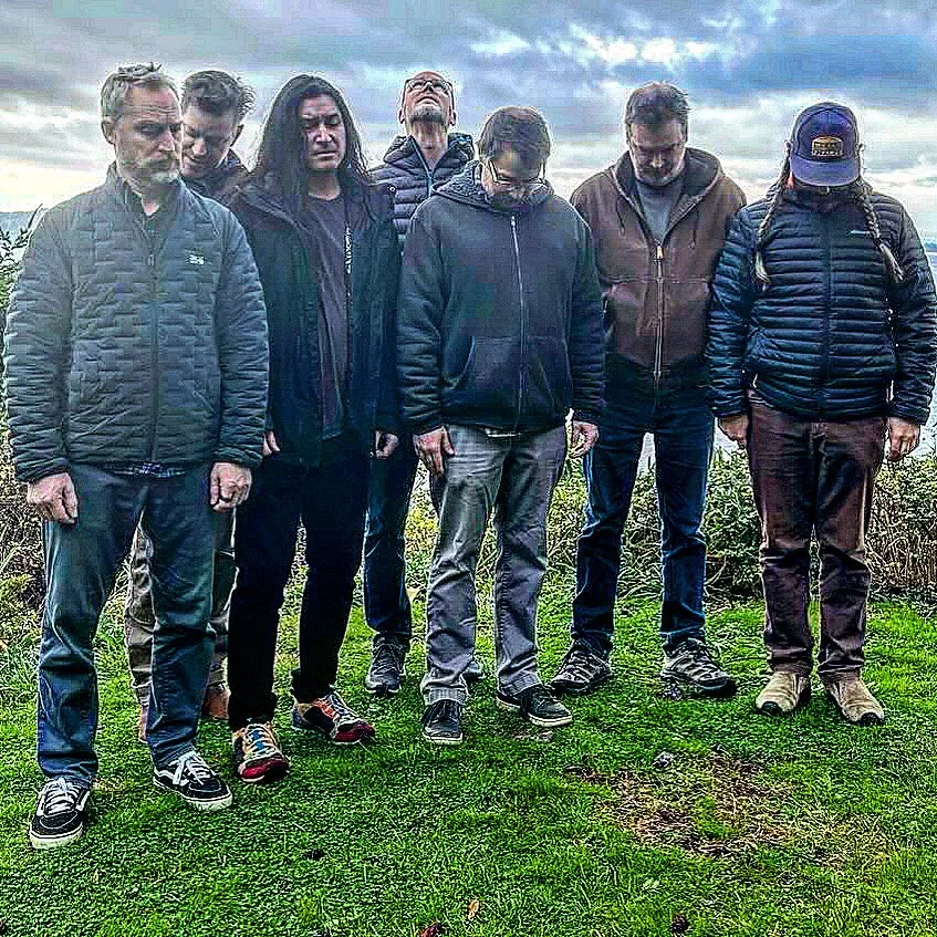Group of six people standing outdoors on grass, some with heads bowed or eyes closed, under cloudy sky.
