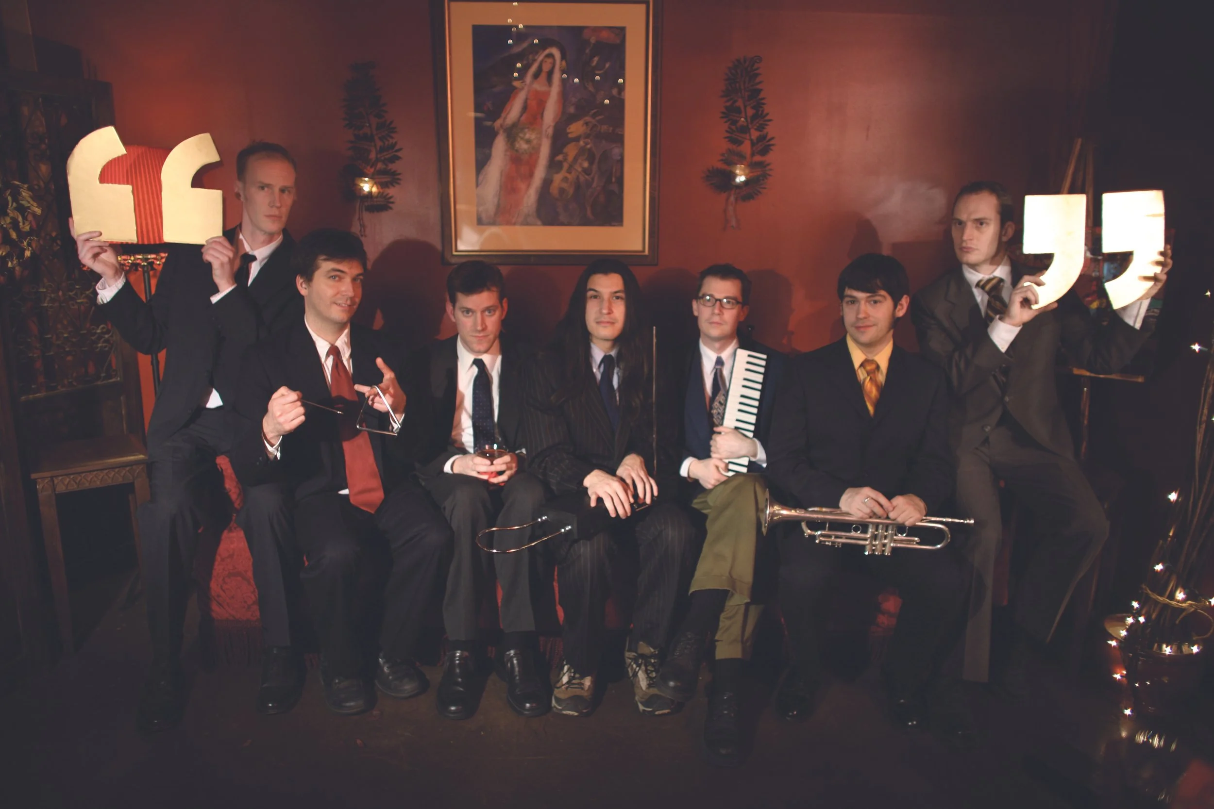 Group of eight men when they were MUCH younger, dressed in suits, sitting and standing in a dimly lit room with a red wall, musical instruments, and a framed painting.