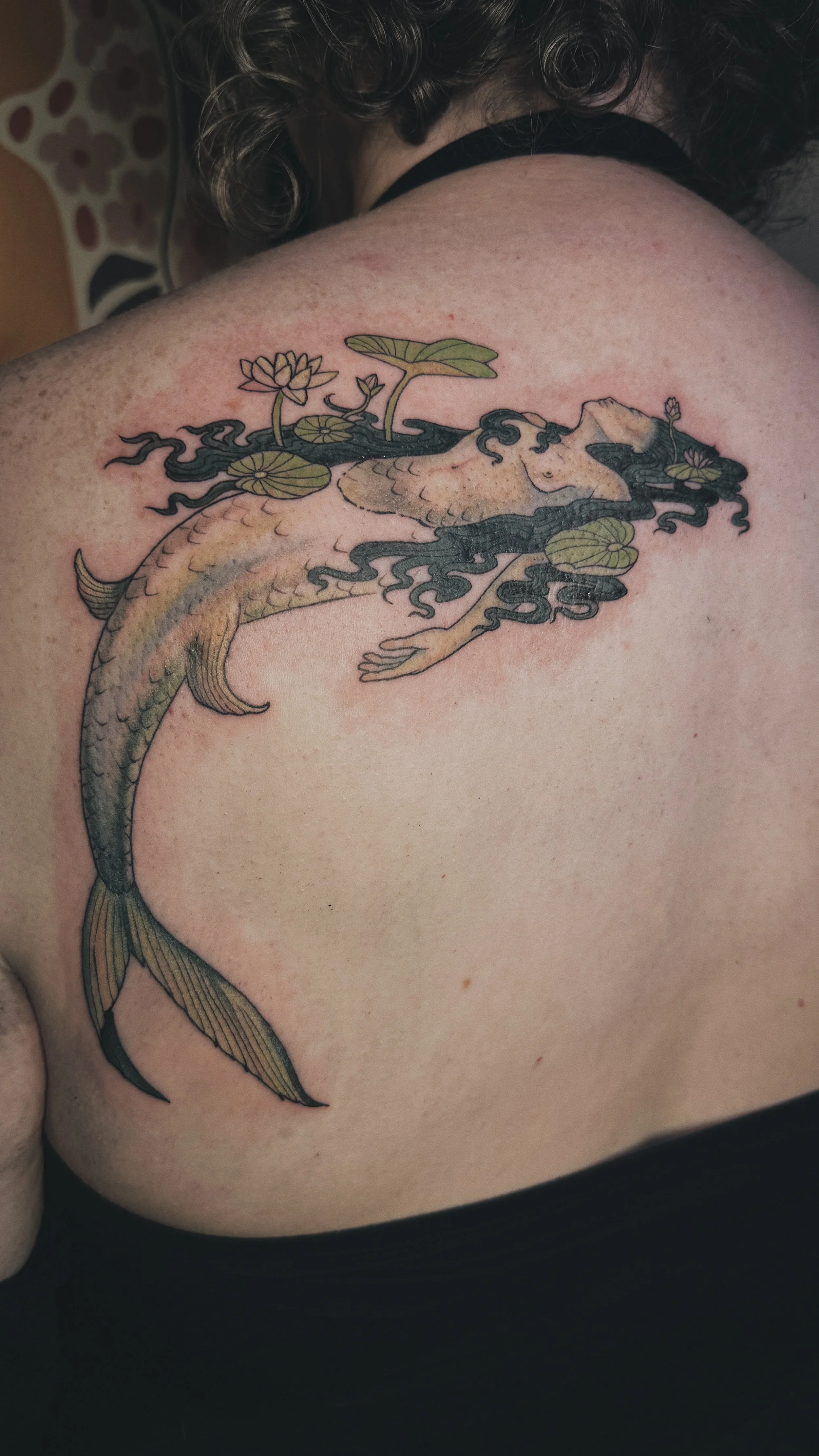 Tattoo of a mermaid with a fish tail, black flowing hair, surrounded by lily pads and lotus flowers on shoulder and upper back.