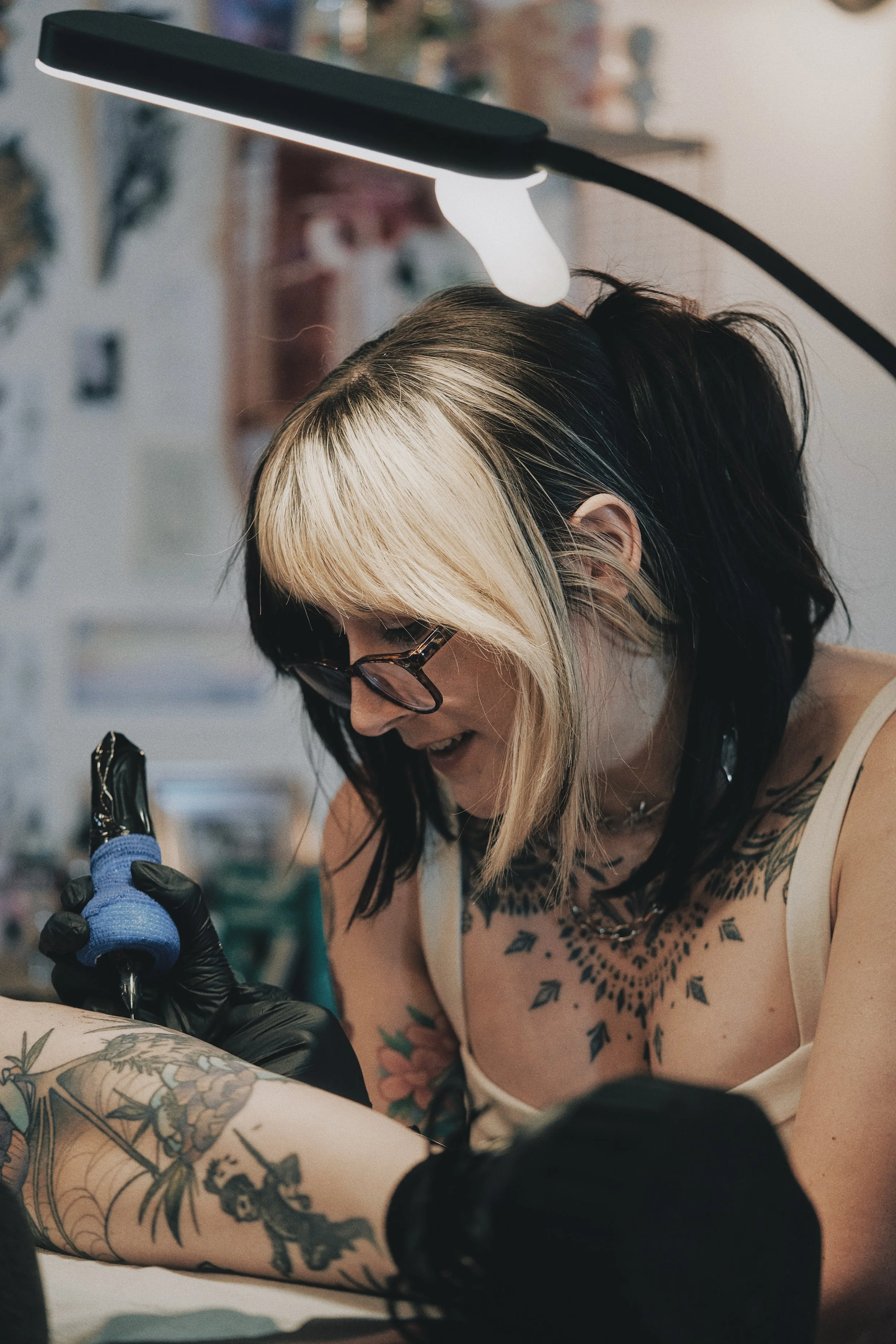 A woman with black and blonde hair, tattoos on her arms and chest, wearing glasses, a necklace, gloves, and a sleeveless top, is tattooing a design on someone's arm under a bright lamp.