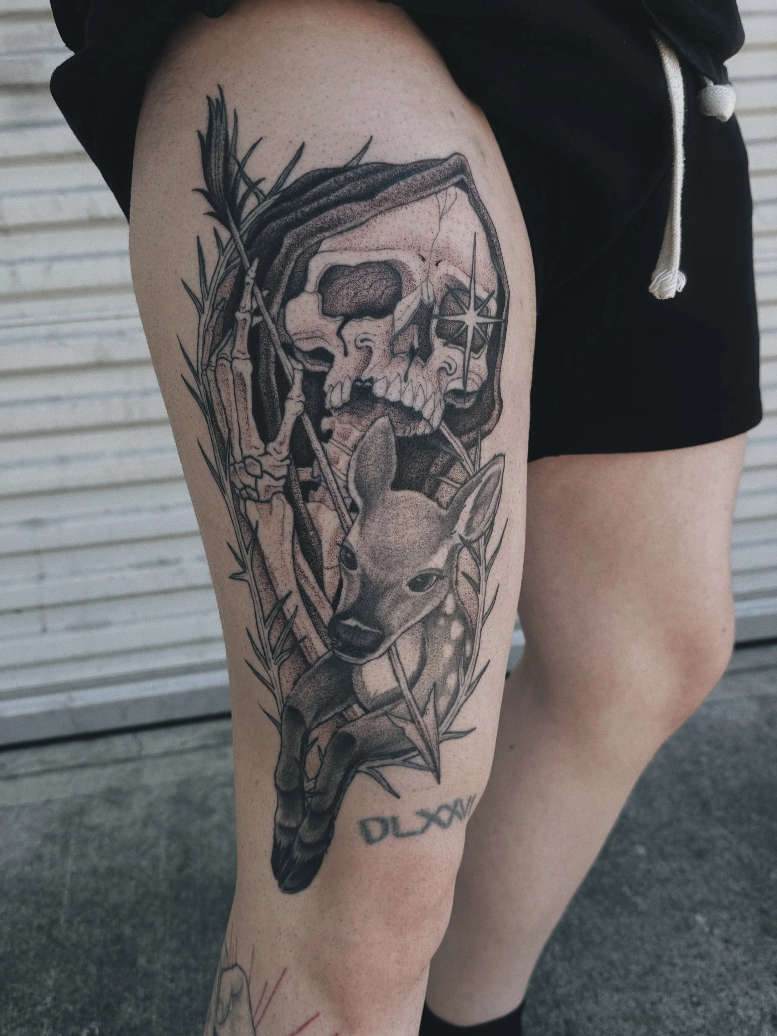 Tattoo of a skull in a cloak holding an arrow , a young deer or fawn, and a star symbol on the skull's eye socket all framed in a thorn boarder on a person's thigh.