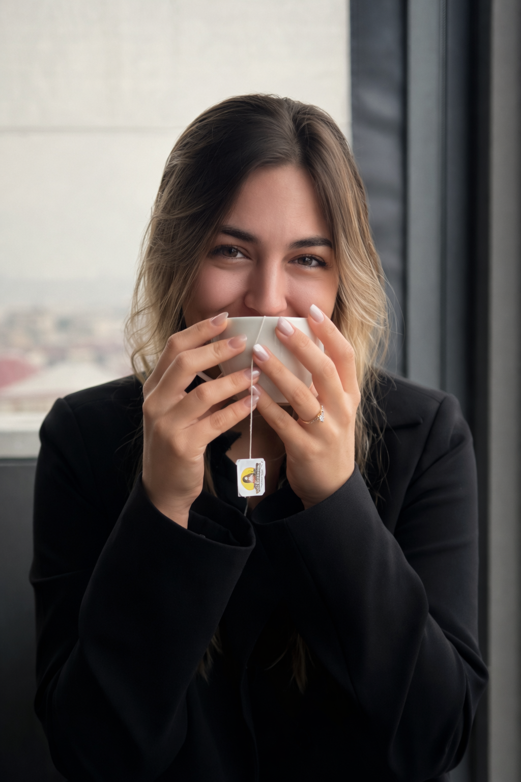 A woman with wavy hair and a black blazer holding a white teacup with a teabag, smiling and partially covering her face behind the cup, sitting by a window in a high-rise building.