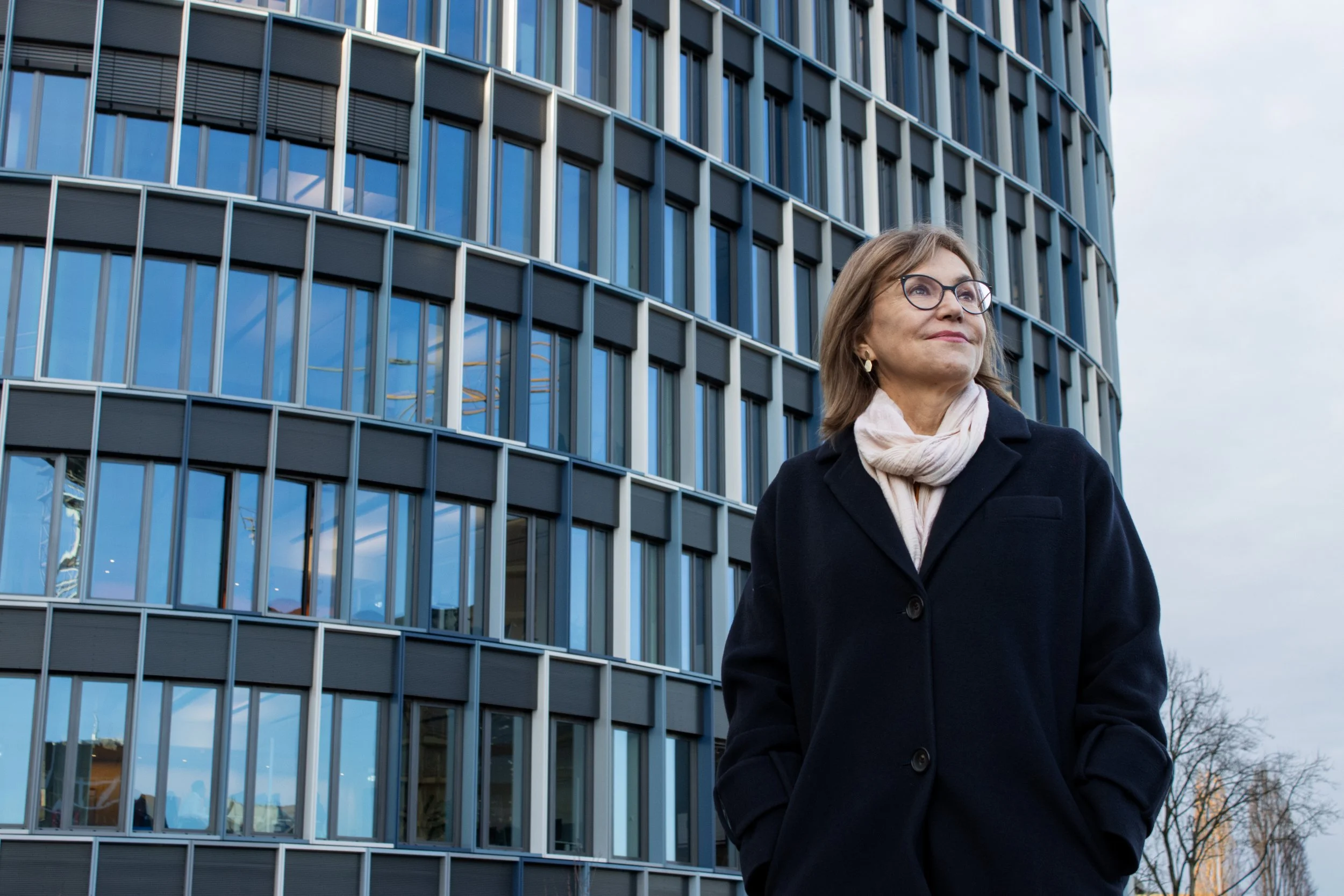 A woman with glasses and a light-colored scarf standing outside a modern glass building with rounded architecture.