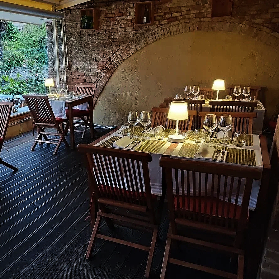Cozy restaurant interior with wooden tables and chairs, set for dining with wine glasses, plates, and cutlery, illuminated by small lamps, brick walls, and large windows showing greenery outside.