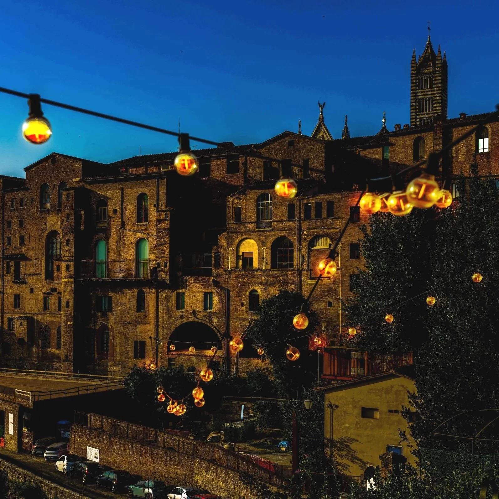 A night scene of an old brick building with string lights hanging in front, glowing warmly against a dark blue sky and silhouetted trees.