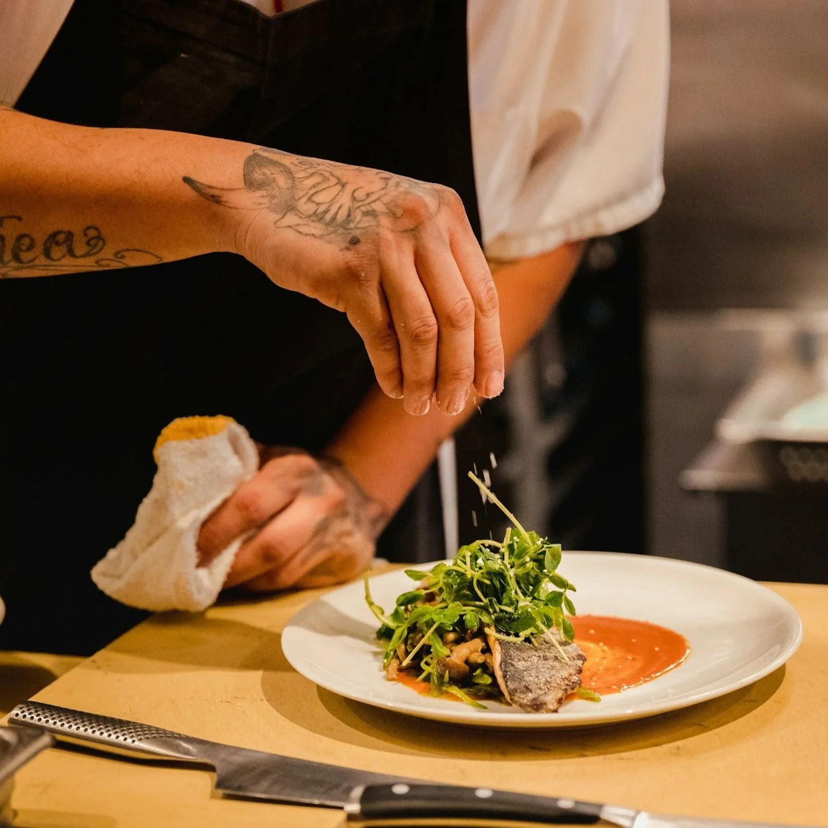 Chef garnishing a plate with fresh greens and fish, using tweezers in a professional kitchen setting.