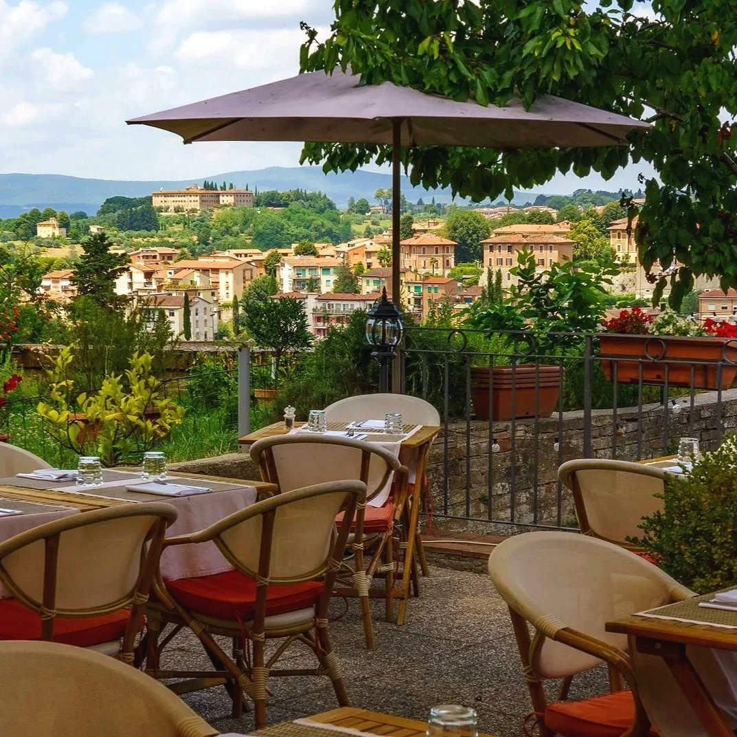 Outdoor restaurant patio with tables, chairs, and an umbrella, overlooking a hillside town with colorful buildings and lush greenery.