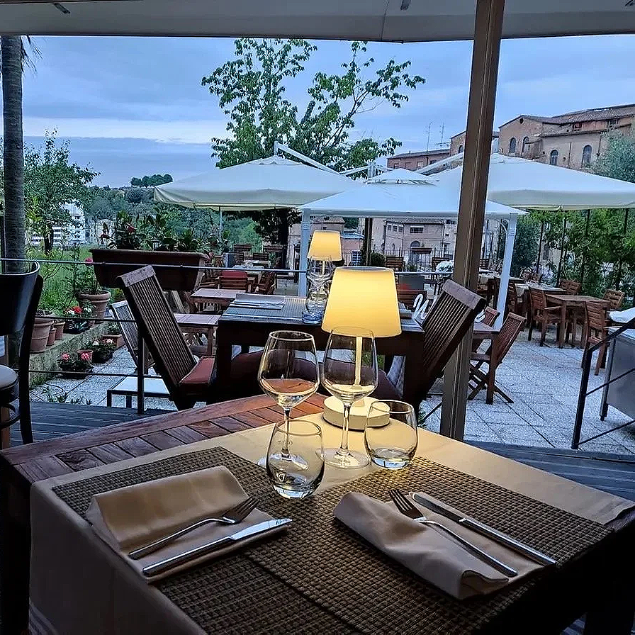Indoor restaurant table set for two with wine glasses, water glasses, napkins, and cutlery, with outdoor patio visible through large windows and illuminated table lamps.