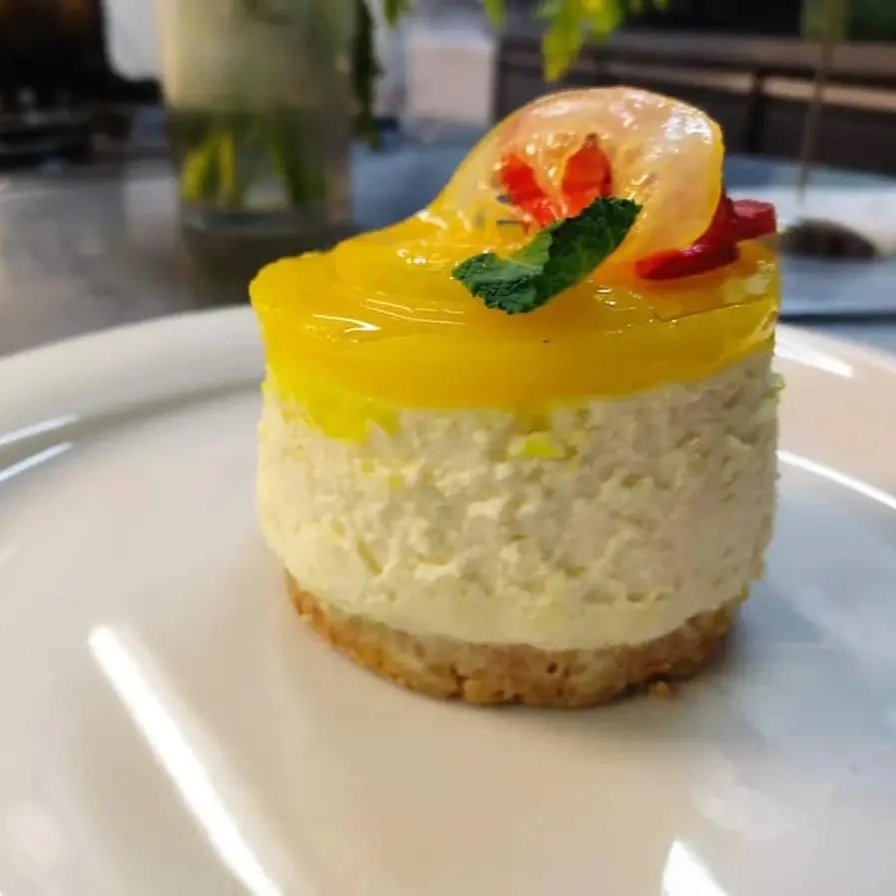 A small, round layered dessert with a biscuit base, creamy middle layer, yellow gelatin top, garnished with a slice of lychee, a mint leaf, and a small red flower petal.