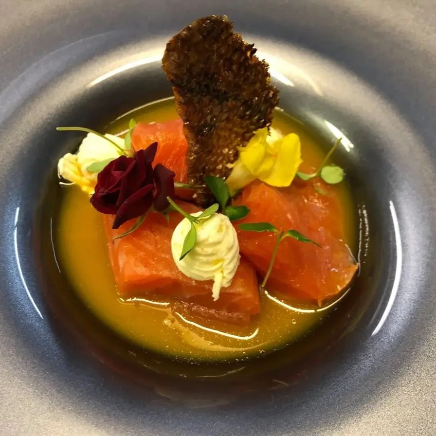 Slices of raw salmon garnished with edible flowers, herbs, and a crispy piece of blackened bread, served in a shallow bowl with a pool of yellow oil or sauce.