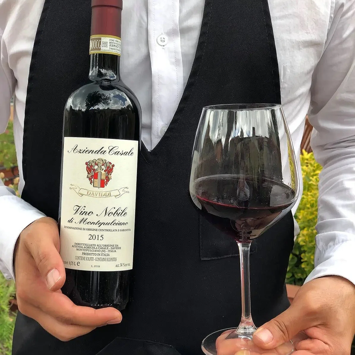 Person wearing a white shirt and black vest holding a bottle of red wine labeled 'Azienda Casale Davidi Vino Nobile di Montepulciano 2015' and a glass filled with red wine, outdoors with greenery and yellow flowers in the background.
