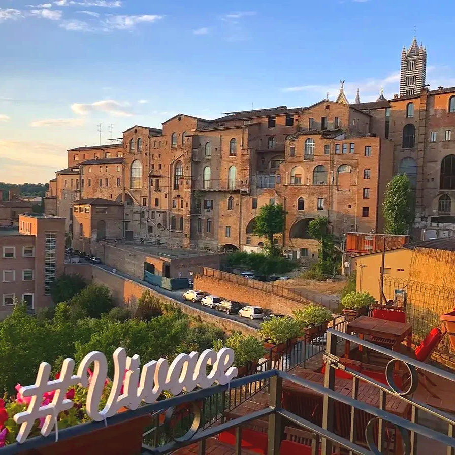 Sunset view of a historic brick building complex with arched windows in a hilly city, seen from a rooftop terrace decorated with plants and outdoor furniture.