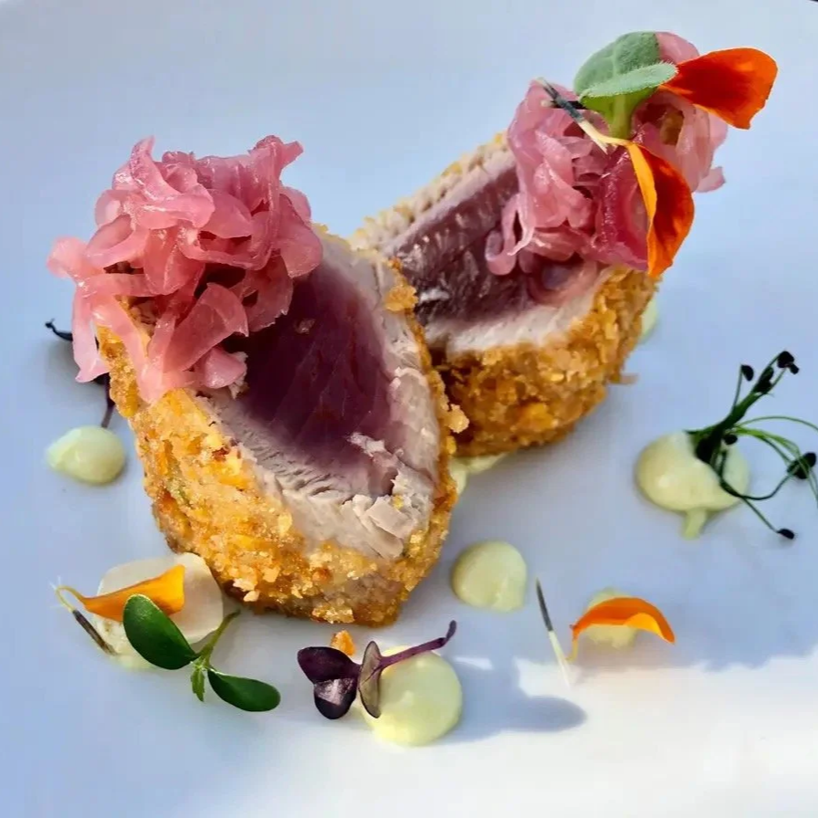 Two slices of breaded, crispy-fried tuna served with pink pickled onions, small dollops of creamy white sauce, and garnished with edible flowers and microgreens on a white plate.