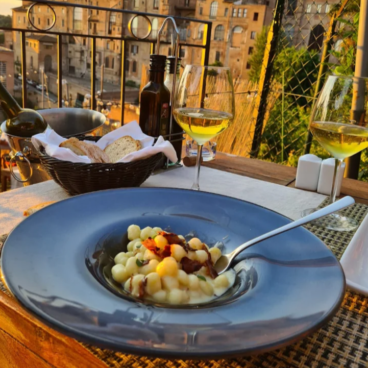 A plate of gnocchi with sauce and toppings on a dining table outdoors with glasses of white wine, bread basket, and city buildings in the background during sunset.