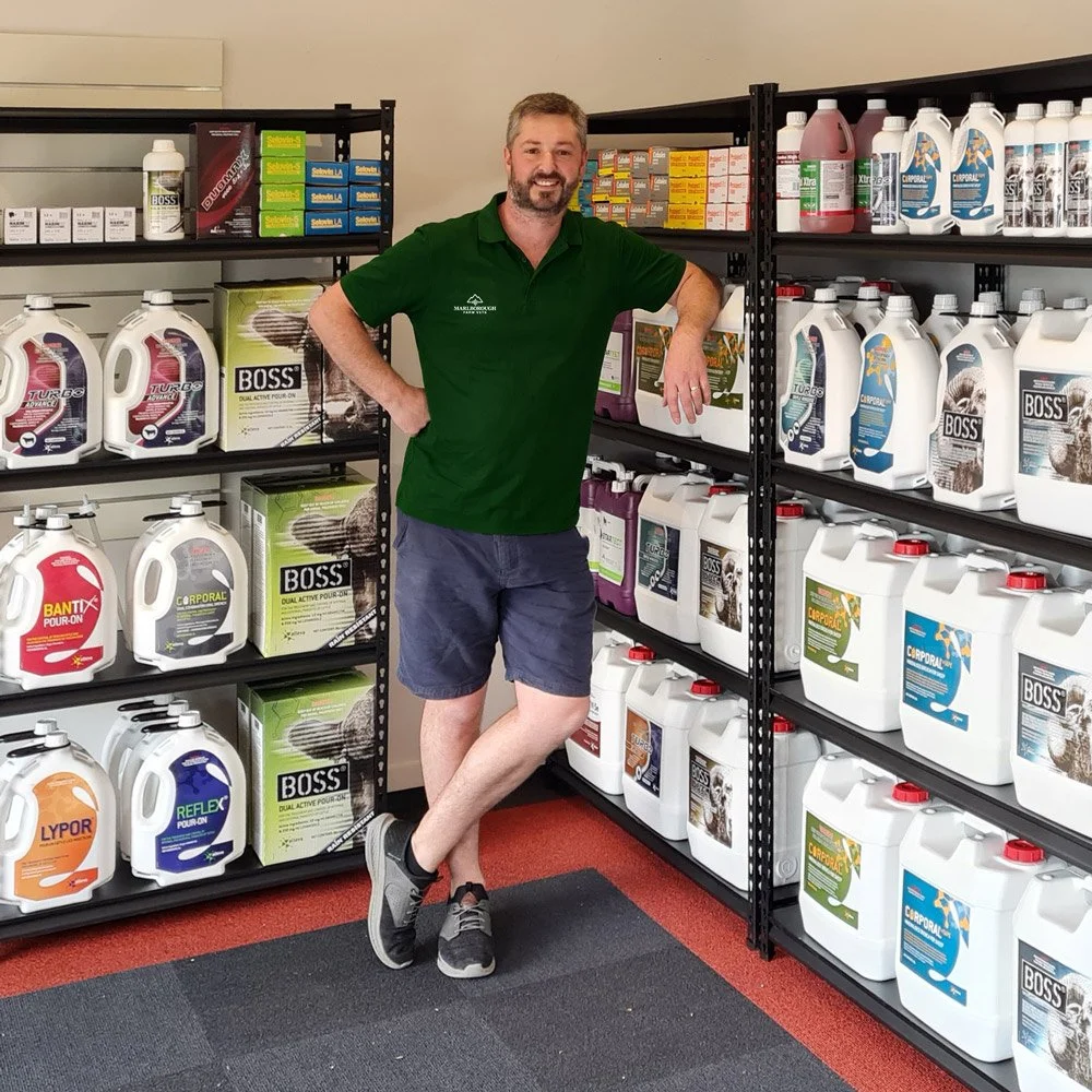 Heath is standing in the Marlborough Farm Vet shop to shelves filled with various bottles of chemicals, supplements,  and supplies.
