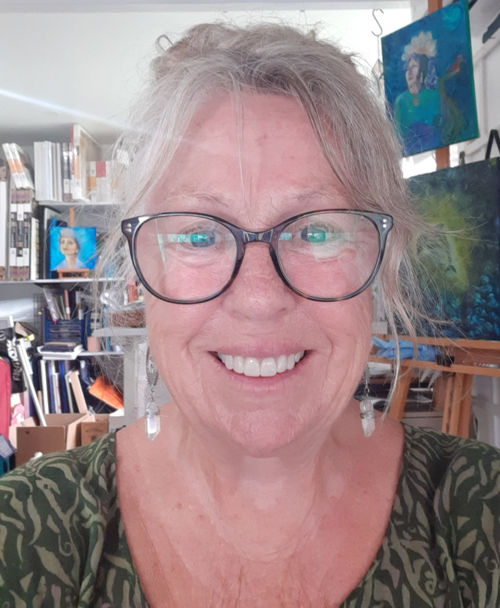 A smiling older woman with gray hair, glasses, and earrings in an art studio.