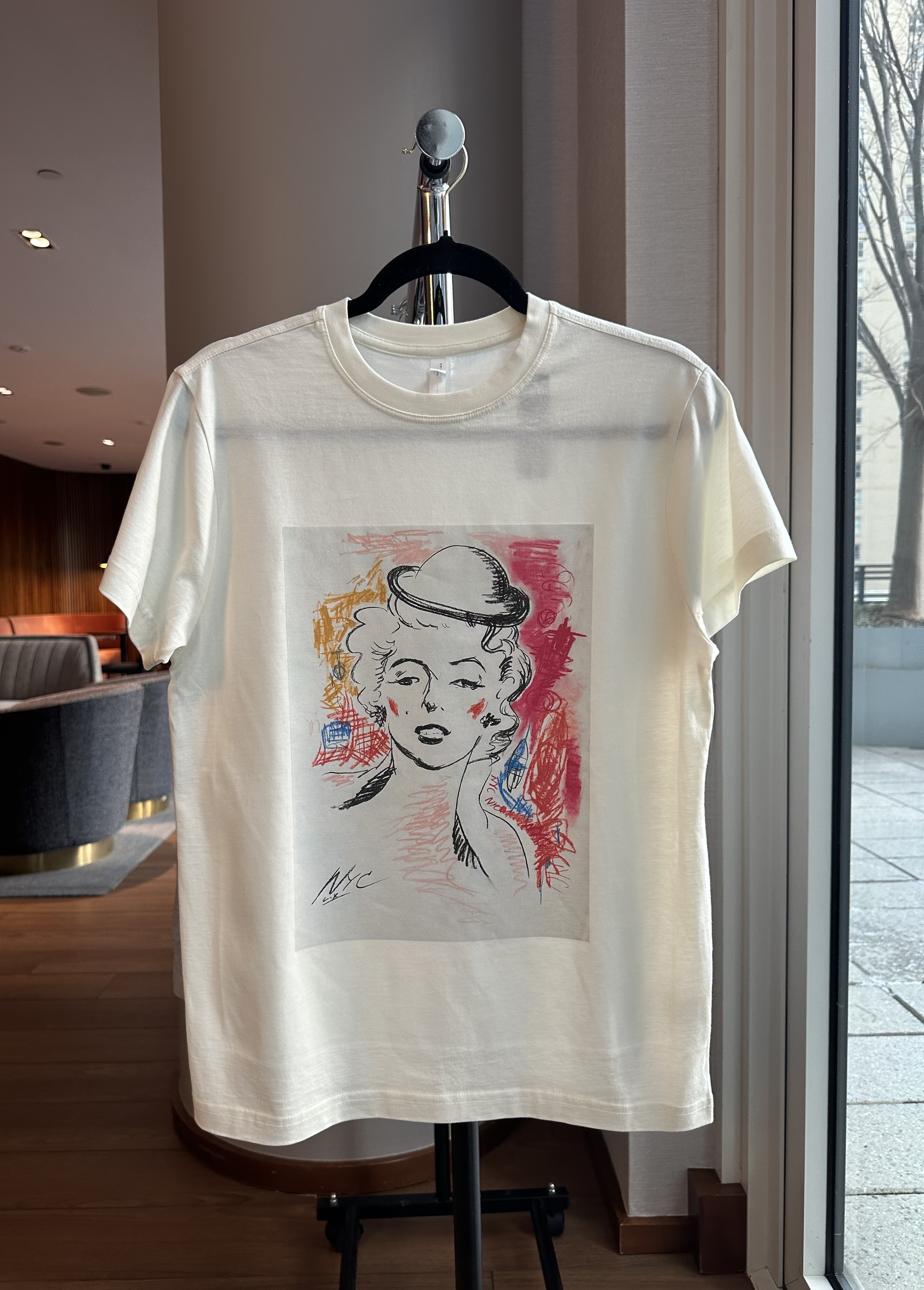 A white T-shirt with a colorful artistic drawing of a woman with curly hair, a hat, and makeup, hanging on a black hanger inside a well-lit indoor space near a glass window.