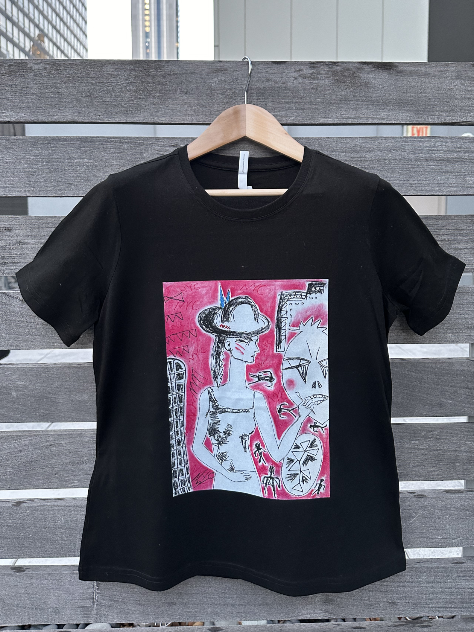Black T-shirt with colorful artistic illustration on the front, hanging on a wooden hanger against a wooden slat background.