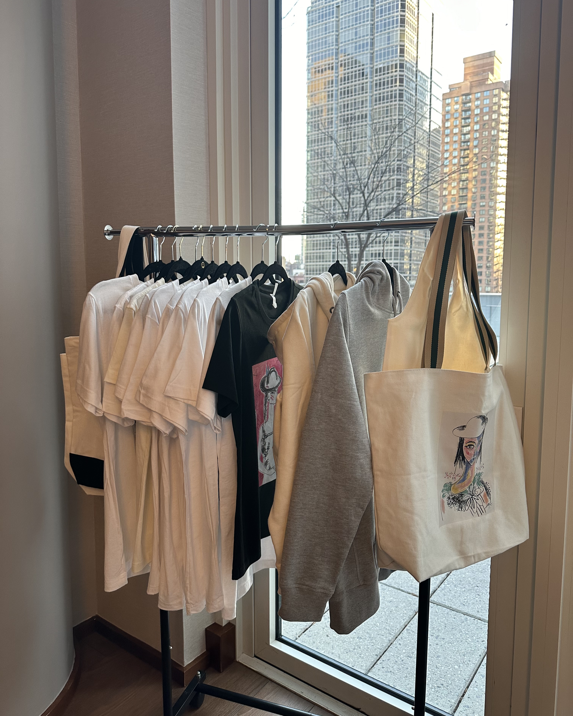 Clothing rack with white, black, and gray shirts and hoodies, displayed near a large window showing tall buildings outside.