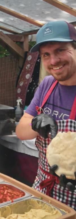 A man wearing a blue cap, purple shirt, and apron is smiling at the camera while holding a food item, possibly a tortilla or flatbread, at an outdoor food stand. The background shows a partially covered outdoor space with wooden beams and a colorful backdrop.