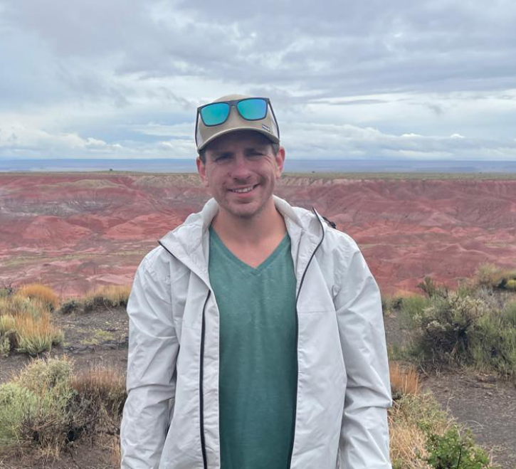 Man standing outdoors in front of colorful canyon with cloudy sky, wearing a white jacket, teal shirt, and sunglasses on cap.