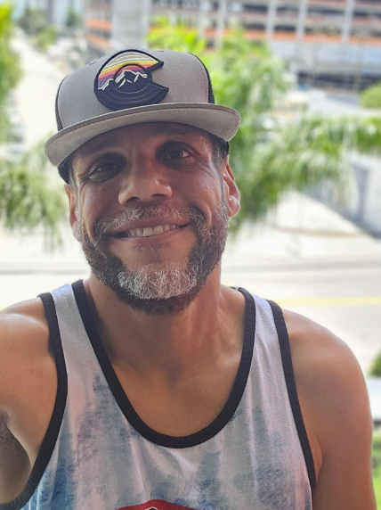 A smiling man outdoors wearing a baseball cap with a mountain and rainbow logo, tank top, and sporting a beard and short hair.